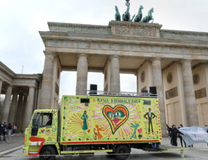 Gelb angestrichener und bunt bemalter Laster steht vor dem Brandenburger Tor in Berlin. In der Mitte des Wagenkastens ist ein Herz und der Titel "Roma Biennale 2020", links ist eine Frauenfigur mit den Namen Papusza gemalt, rechts eine Männerfigur von Helios Gomez